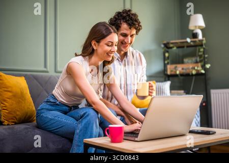 Adult couple is sitting at sofa at home and using laptop. They are shopping online and searching for goods. Stock Photo