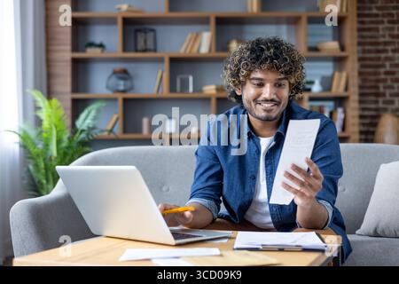 A cheerful man sits on a couch at home, using his laptop while examining a piece of paper. The atmosphere is casual and relaxed, suggesting productivi Stock Photo