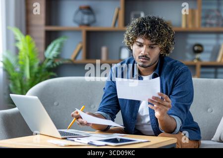 A thoughtful man examines financial papers while working on a laptop in a stylish home office. The setting exudes modernity and concentration, reflect Stock Photo