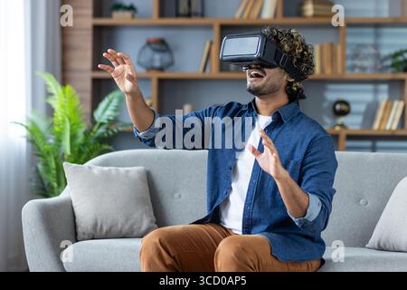 A man wearing a VR headset is exploring a virtual experience while seated on a sofa. The background features a neatly arranged bookshelf and indoor pl Stock Photo