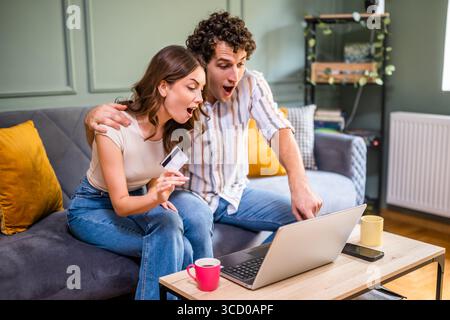 Adult couple is sitting at sofa at home and using laptop. They are shopping online. Woman is holding credit card. Stock Photo