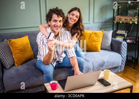 Adult couple is sitting at sofa at home and using laptop. They are shopping online. Man is holding credit card. Stock Photo