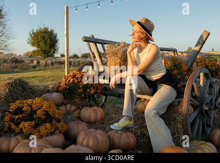 Happy woman sitting on wooden cart near many pumpkins in park on autumn day. Stock Photo