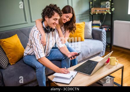 Adult couple is sitting at sofa at home and using laptop.  They are shopping online and making shopping list. Stock Photo