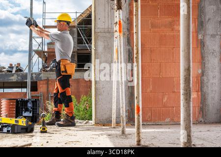 Caucasian construction worker is carefully measuring a wall using a level tool at a busy construction site during daylight hours, surrounded by scaffo Stock Photo
