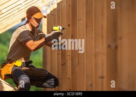 A skilled carpenter is working diligently to secure wooden boards using a power drill on a sunny day at a construction site surrounded by greenery. Stock Photo