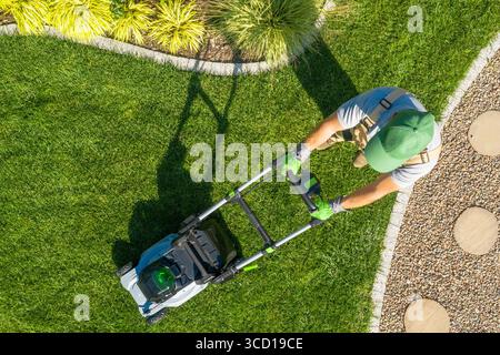 A lawn care professional trims lush green grass in a neatly landscaped garden during daylight. Freshly cut grass lines contrast with stone pathway. Stock Photo