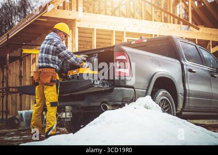 A construction worker in yellow gear prepares tools from a truck at a snowy building site. Wooden frames are visible, indicating an active constructio Stock Photo