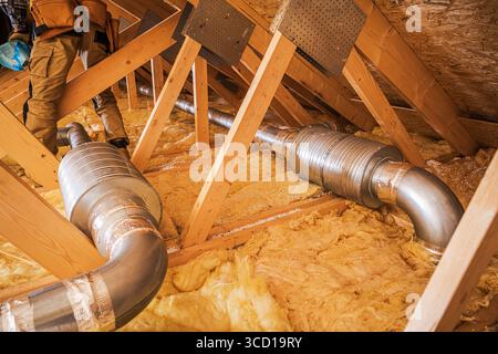 Workers are installing metal ductwork in an attic filled with insulation material. Proper ventilation is crucial for energy efficiency and air quality Stock Photo