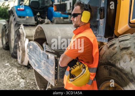 A construction worker stands beside heavy machinery, wearing safety gear and sunglasses, resting briefly during a busy workday at a construction site. Stock Photo
