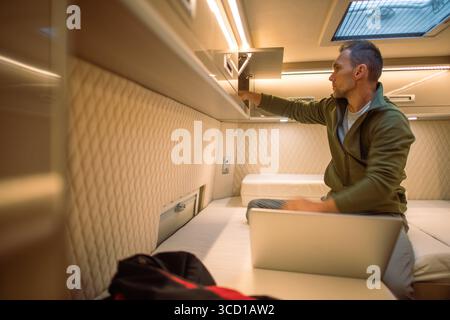 A man sits in a stylish camper van, focused on his laptop while organizing storage. Warm lights illuminate the cozy space during evening hours. Stock Photo