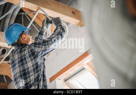 An electrician installs wiring in the ceiling of a home under renovation. He is focused on his task while wearing safety gear and working carefully. Stock Photo