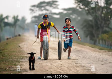 Bogura, Bangladesh - 13 December 2019: View of two boys racing with tires down a dirt path as a small goat looks on, the cool tones of the sky contrasting with the warm earth tones of the path. Stock Photo