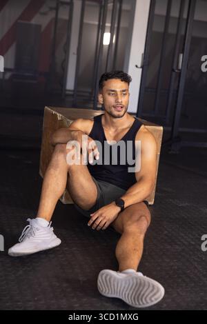 Asian man sitting on gym floor leaning against plyometric box wearing sportswear with squat racks Stock Photo