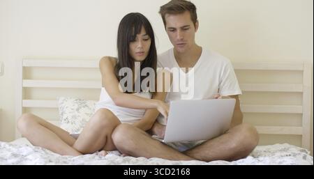 Smiling young long haired woman showing something to handsome man on laptop while in bed Stock Photo
