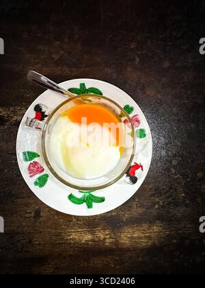 A glass cup with half-boiled eggs on a decorative plate with a spoon, placed on a dark textured table cafe penang, malaysia - kopitiam eggs Stock Photo