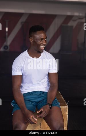 African American man sitting on wooden plyometric box at gym, holding smartwatch and smiling Stock Photo