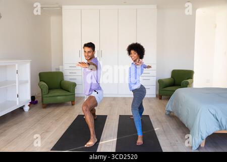Diverse friends performing warrior II poses on black yoga mats in bright home exercise room Stock Photo