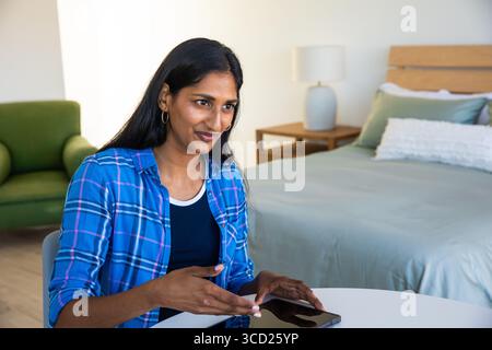 Indian woman sitting at round white table near bed, using smartphone in bright bedroom, copy space Stock Photo