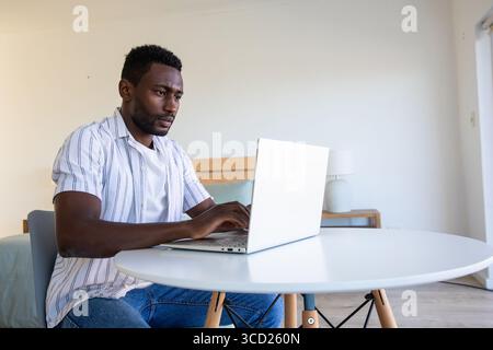 African American man typing on silver laptop at round white table in minimalist bedroom, copy space Stock Photo