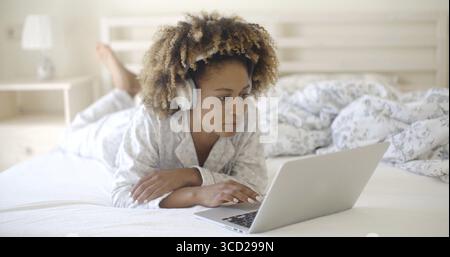 Woman lying on her bed while using a laptop and listening to headphones and smiling Stock Photo