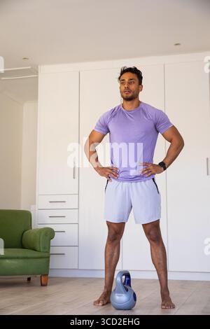 Asian man standing barefoot on wood floor in home workout space with grey kettlebell Stock Photo