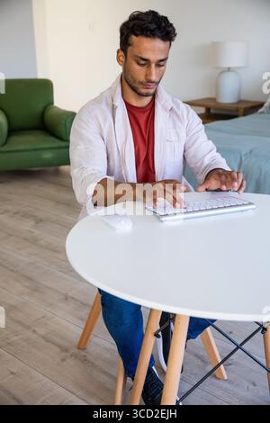 Man typing on wireless keyboard at small round table in bedroom living area with wireless mouse Stock Photo