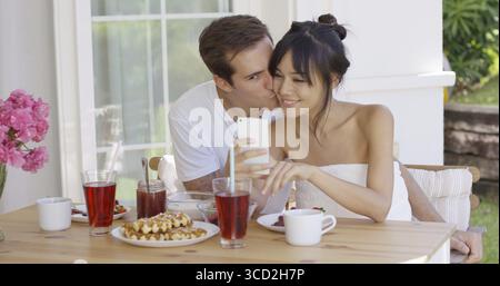 Happy Mixed-race couple sitting on terrace having a teasty breakfast. They taking selfie photo with their mobile phone Stock Photo