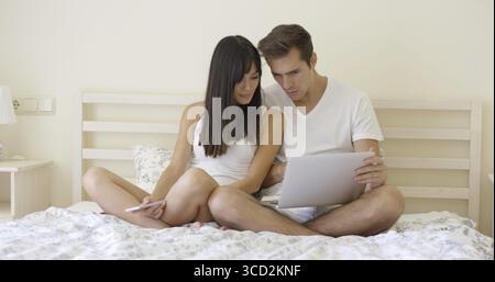 Young couple sitting together in underclothes while on bed while doing research on a laptop computer and smart phone Stock Photo