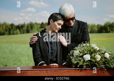Middle aged Caucasian woman leaning on senior Caucasian man wearing sunglasses standing beside wooden coffin with floral arrangement outdoors during funeral ceremony Stock Photo