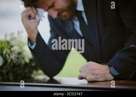 Middle aged Caucasian man leaning over coffin wiping tears with tissue during funeral ceremony, showing visible grief and emotional distress, blurred background with greenery Stock Photo