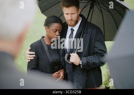 Caucasian middle aged man comforting Black young adult woman under umbrella during funeral ceremony outdoors, both standing close together with solemn expressions Stock Photo