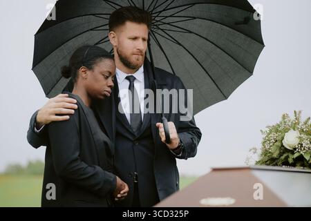 Middle aged Caucasian man comforting middle aged Black woman under umbrella standing beside coffin during funeral ceremony outdoors, both showing somber expressions Stock Photo
