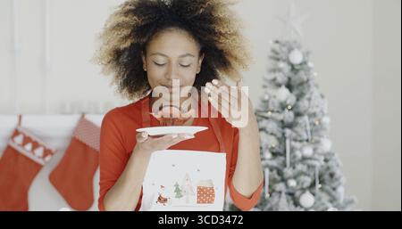 Young African cook with a freshly baked Christmas cake in her hand closing her eyes in bliss as she enjoys the aroma Christmas tree background Stock Photo