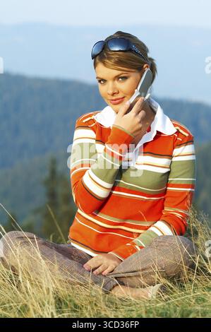Young beautiful woman relaxing on meadow at mouintain outdoors and using cell phone Stock Photo