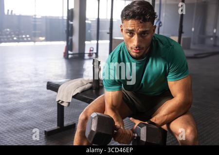 Muscular man lifting dumbbell on gym bench amid racks and kettlebells, using towel, water bottle Stock Photo