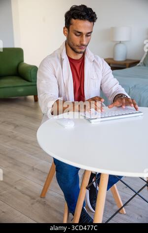 Man typing on wireless keyboard at round white table in bedroom workspace with mouse and lamp Stock Photo