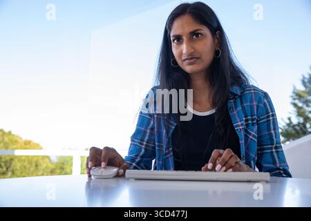 Indian woman typing on white keyboard and using wireless mouse at balcony desk with glass railing Stock Photo