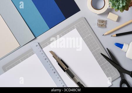 White sheet with ruler and knife on cutting mat among craft tools and colored paper. Creative workspace for art and design preparation. Stock Photo
