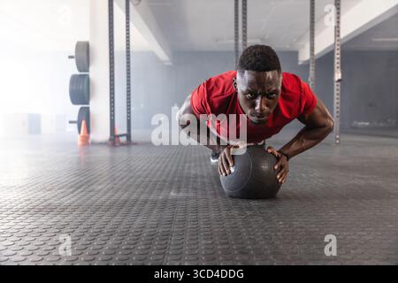 African American man in sportswear doing pushups on medicine ball in gym near orange cones Stock Photo