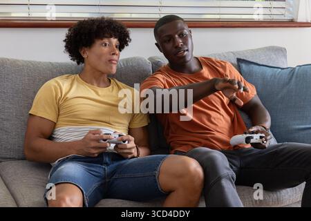 Diverse male friends sitting on living room sofa by blinds holding white gamepads with blue cushion Stock Photo