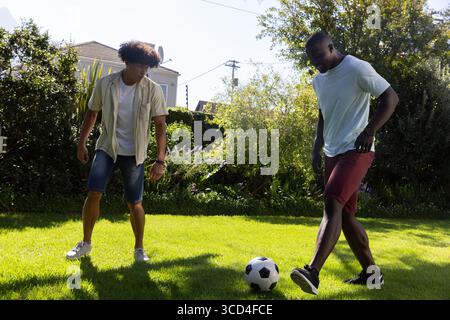 Diverse male friends wearing shorts playing soccer on green lawn in yard with black-and-white ball Stock Photo