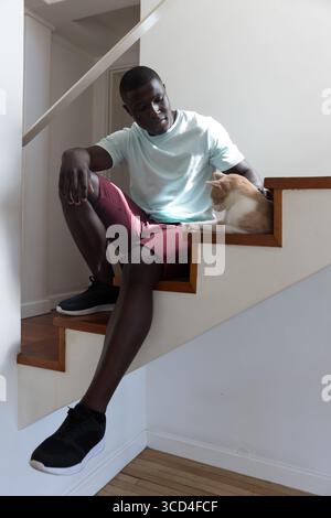 African American man sitting on wooden staircase inside home petting ginger and white cat Stock Photo