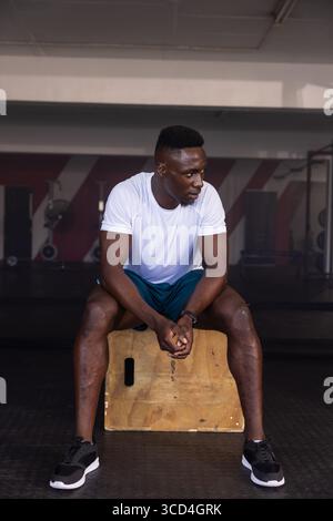 African American male resting on plyometric box at gym beside weight machines and rubber matting Stock Photo