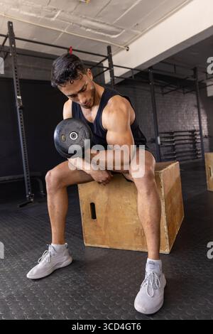 Man doing concentration curl while sitting on wooden box at gym wearing sportswear with dumbbell Stock Photo