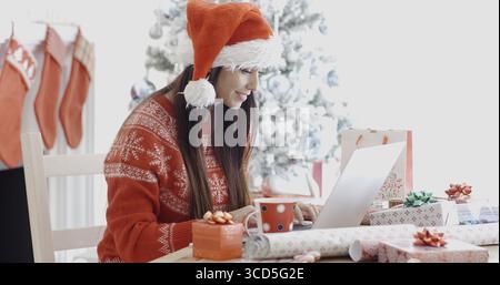 Smiling young woman surfing the internet on her laptop computer for Christmas bargains as she sits at a table in front of the decorated tree in her re Stock Photo