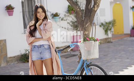 A joyful woman standing next to a blue bicycle on a cobblestone street in Spain, exuding happiness and casual style Stock Photo