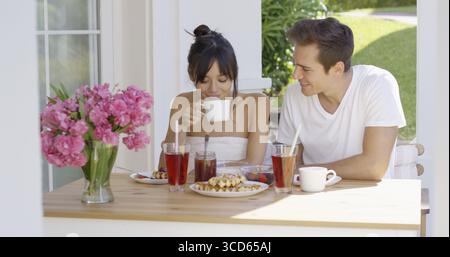 Couple having breakfast at outdoor table with pink flowers in vase and green grass in background Stock Photo