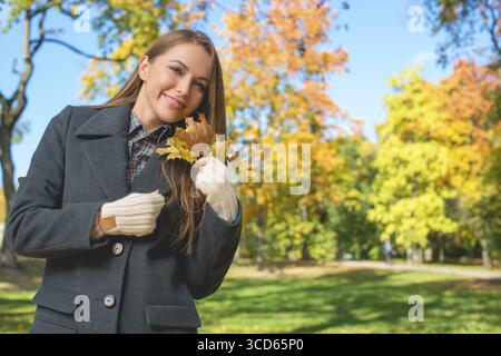 Smiling Young Blond Woman in Gray Coat Holding Dry Leaves. Captured Outdoor with Nature Background Stock Photo