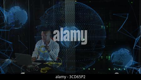Engineer wearing tie plugging yellow Ethernet cables at server room desk, with network globes Stock Photo
